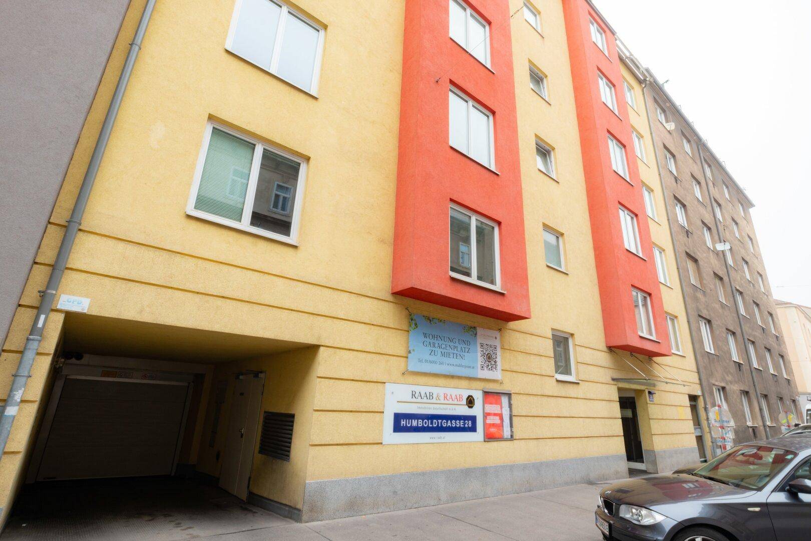 Fassade eines modernen Mehrfamilienhauses mit gelben und roten Akzenten sowie Garageneinfahrt.