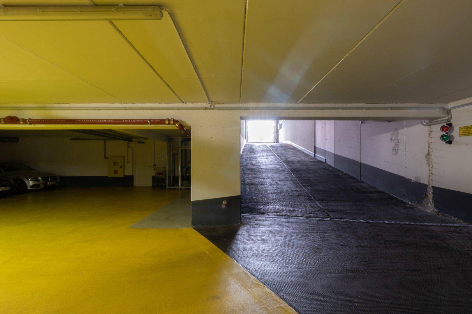 Einfahrt zur Tiefgarage mit Rampe, gelbem Bodenbereich und Licht am Ausgang.