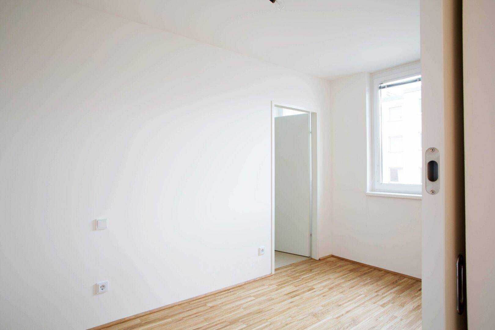 Helles Schlafzimmer mit Parkettboden und Fenster, bereit zur individuellen Gestaltung.