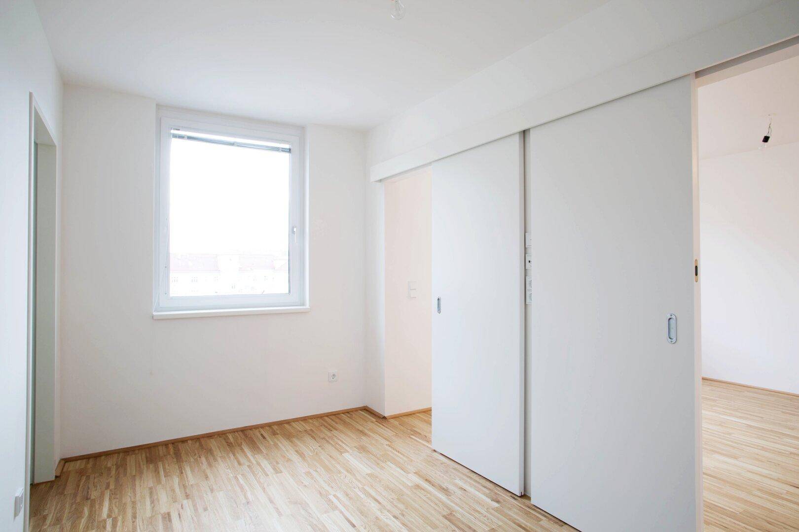 Geräumiges Schlafzimmer mit Parkettboden, großem Fenster und praktischer Schiebetür.