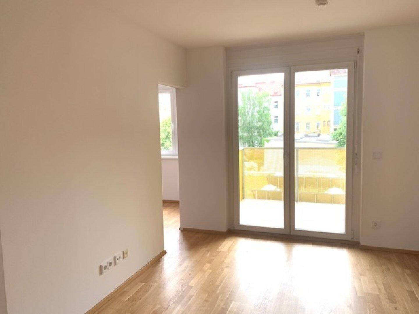 Lichtdurchfluteter Wohnraum mit Holzboden und großen Fenstern, die auf einen Balkon führen.