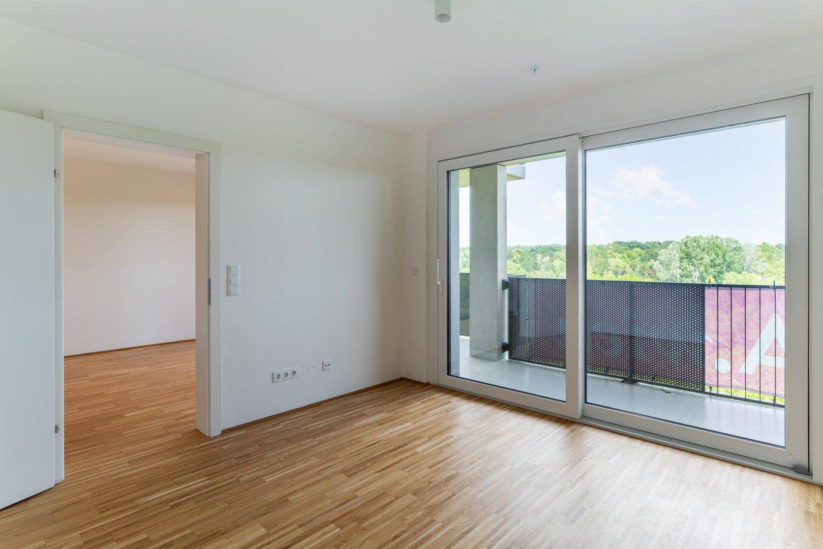 Helles Schlafzimmer mit Parkettboden und großem Schiebefenster zum Balkon mit grüner Aussicht.