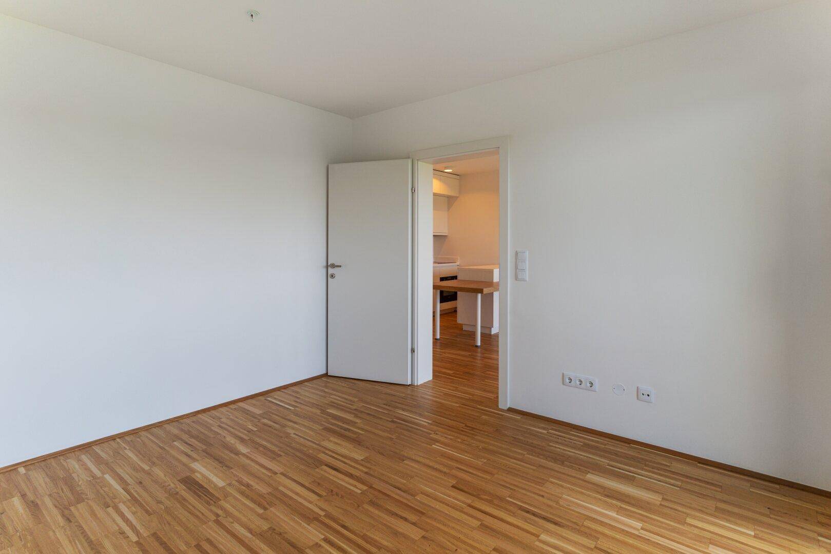 Leerer Raum mit Parkettboden und Zugang zur modernen Küchenzeile der Wohnung.