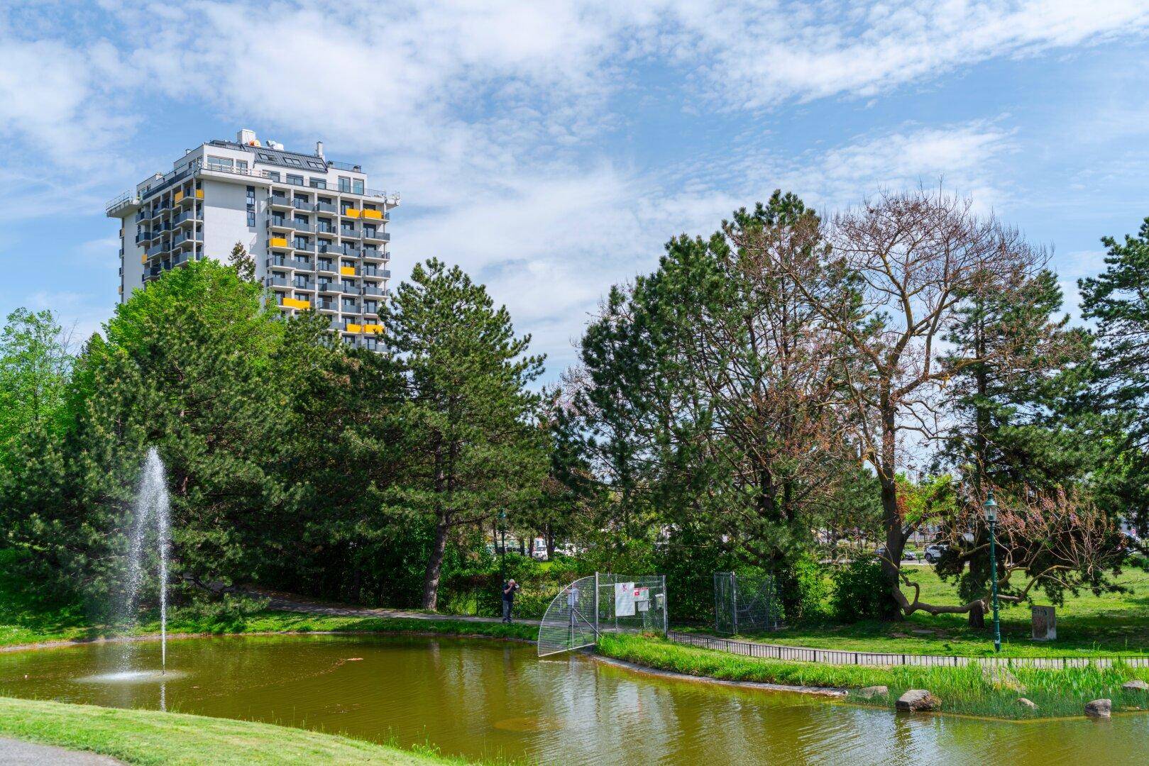 Hohes Wohngebäude mit Balkonen, umgeben von einem Park mit Teich und Springbrunnen.
