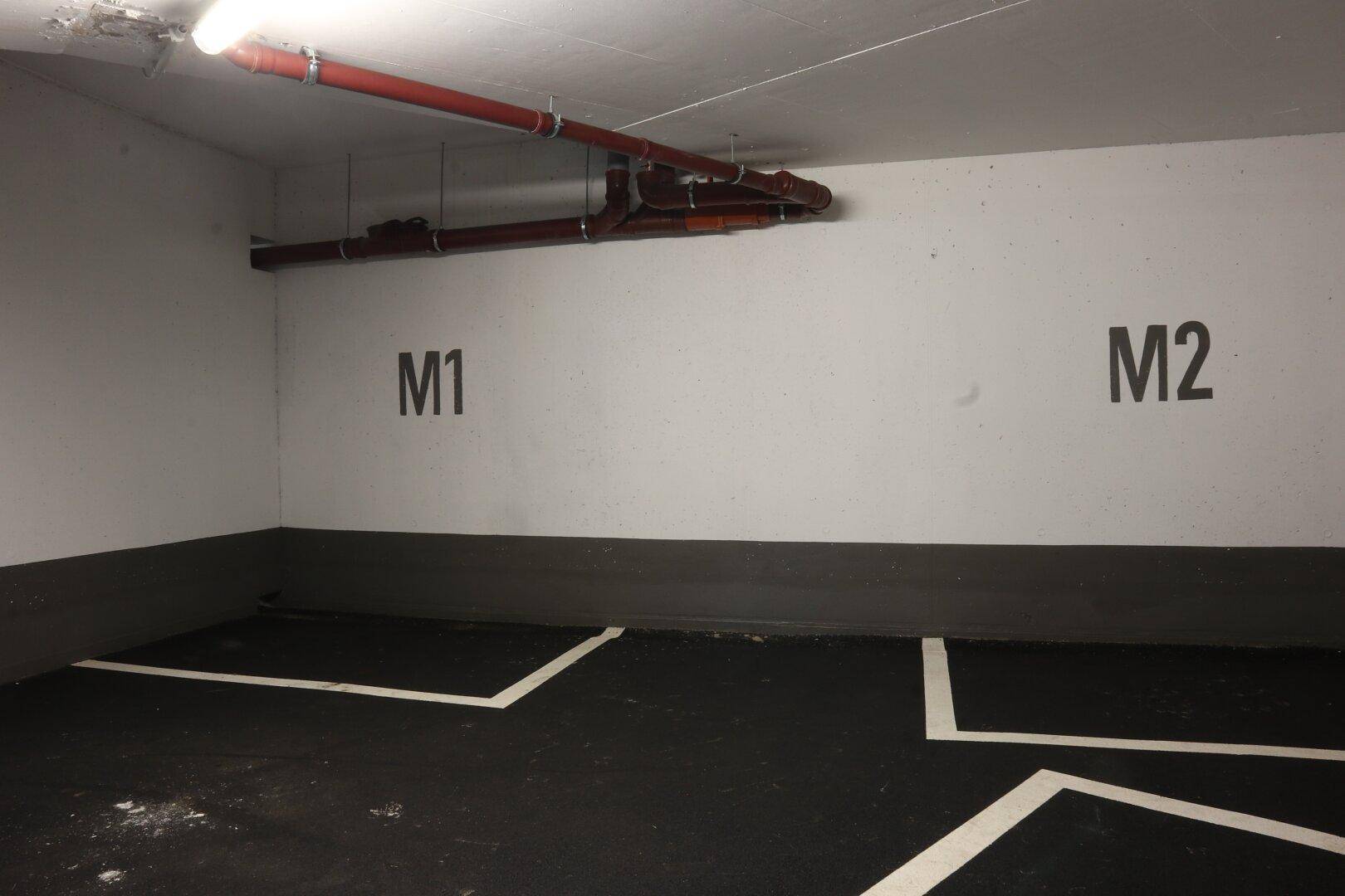 Saubere Tiefgarage mit Stellplätzen M1 und M2, roten Rohren an der Decke und heller Beleuchtung.