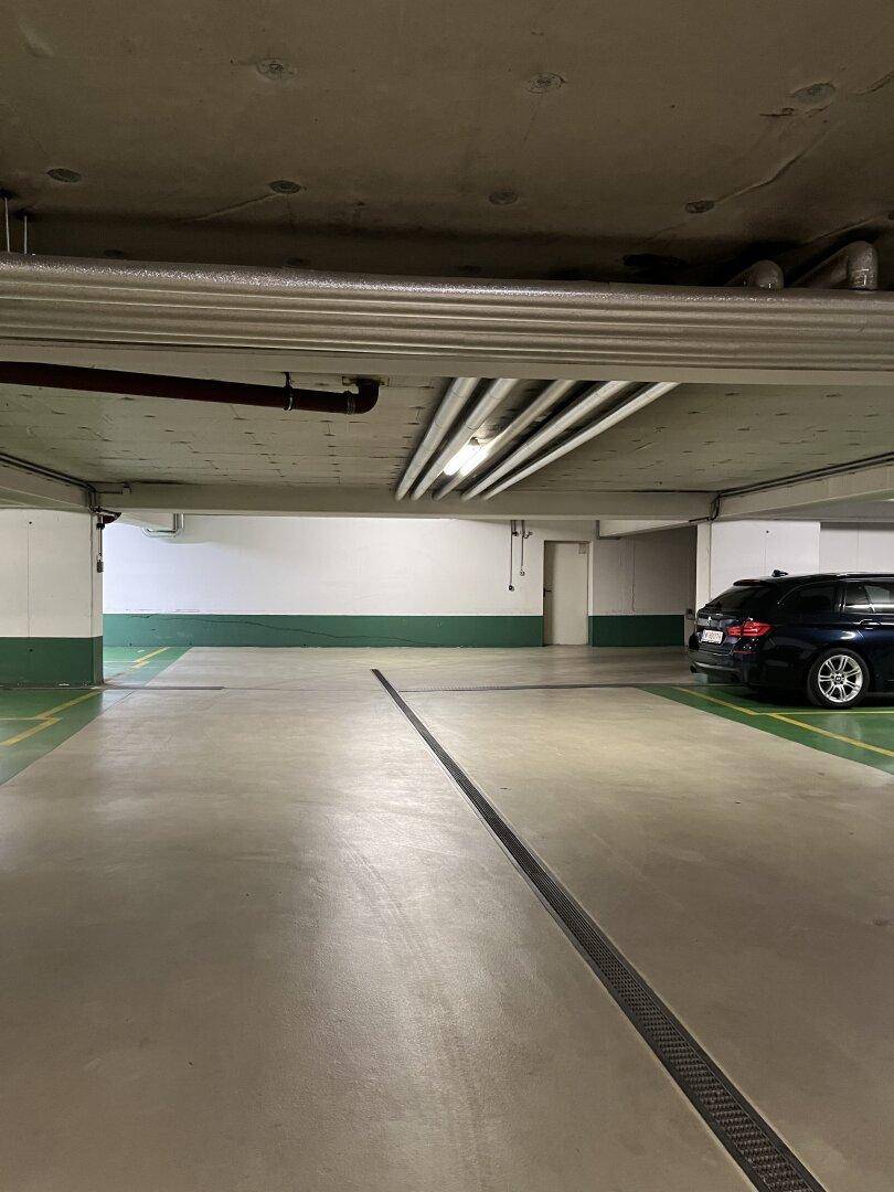 Leere Tiefgarage mit sichtbaren Leitungen an der Decke und einem geparkten Auto.