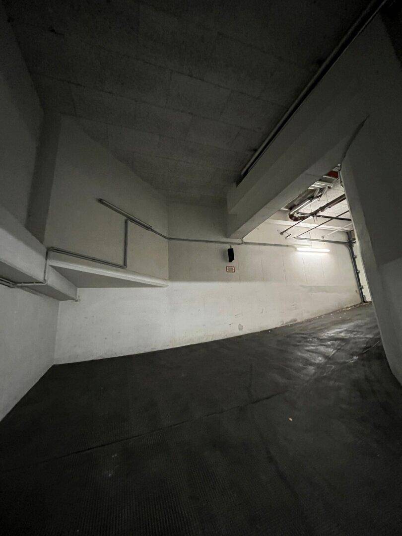 Dunkle Tiefgarage mit einer geschwungenen Rampe und sichtbaren Leitungen an der Decke, beleuchtet von einzelnen Lampen.