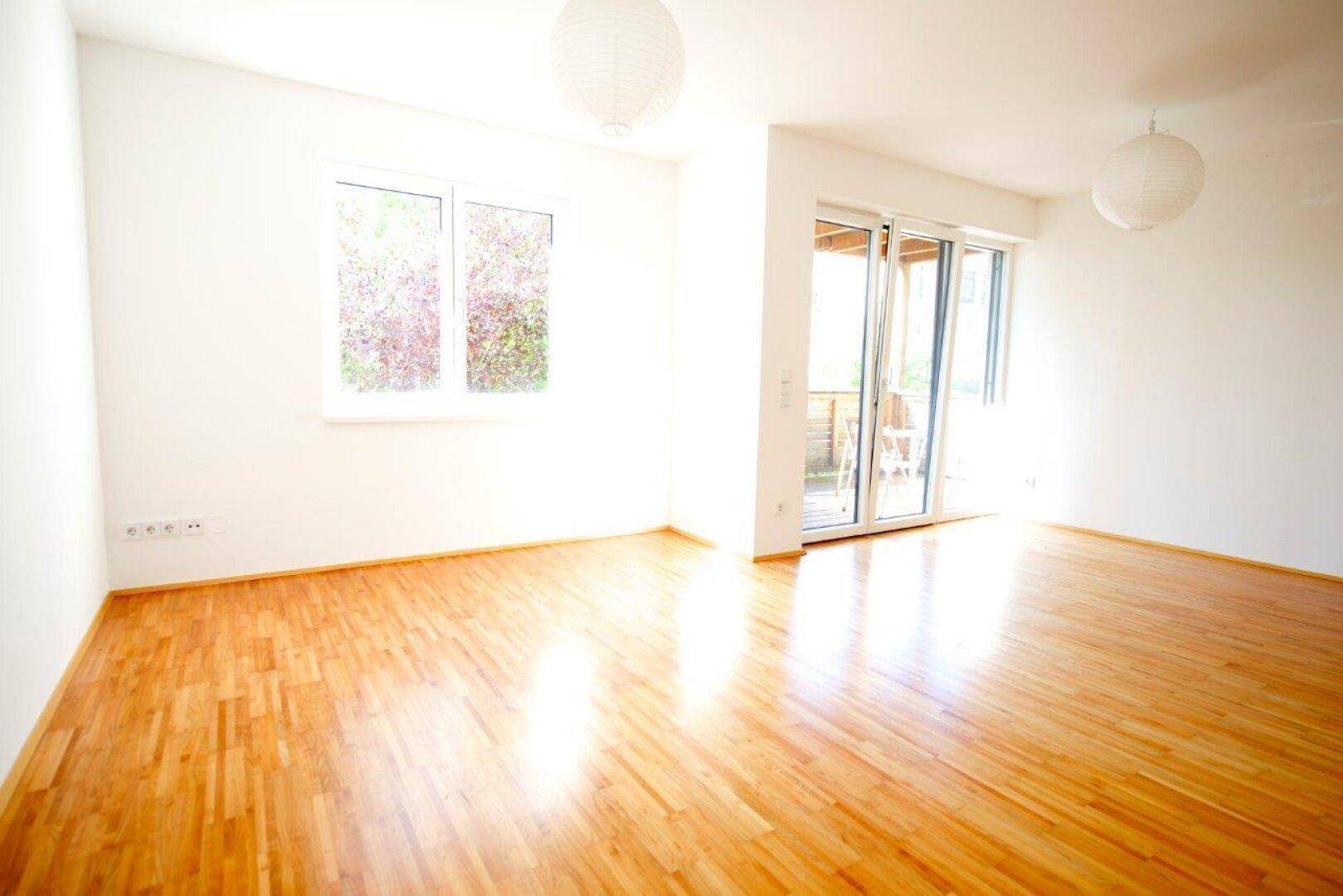 Heller Wohnraum mit Parkettboden, großen Fenstern und Zugang zum Balkon.