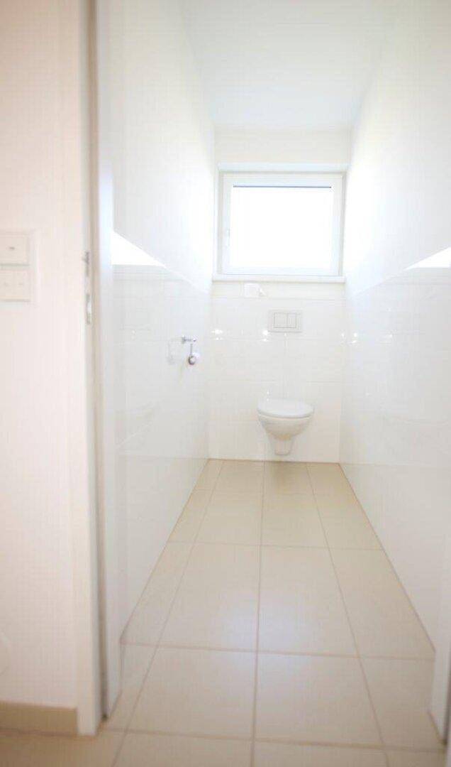 Schlichtes Gäste-WC mit Wand-WC, Fliesenboden und Fenster für Tageslicht.