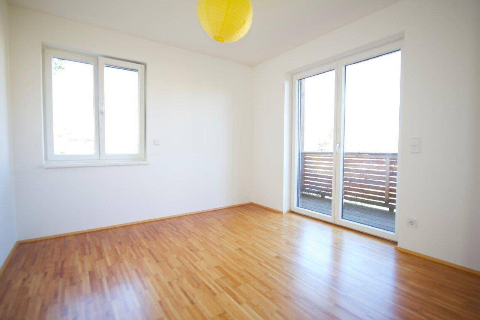 Schlafzimmer mit Parkettboden, großem Fenster und Zugang zum Balkon.