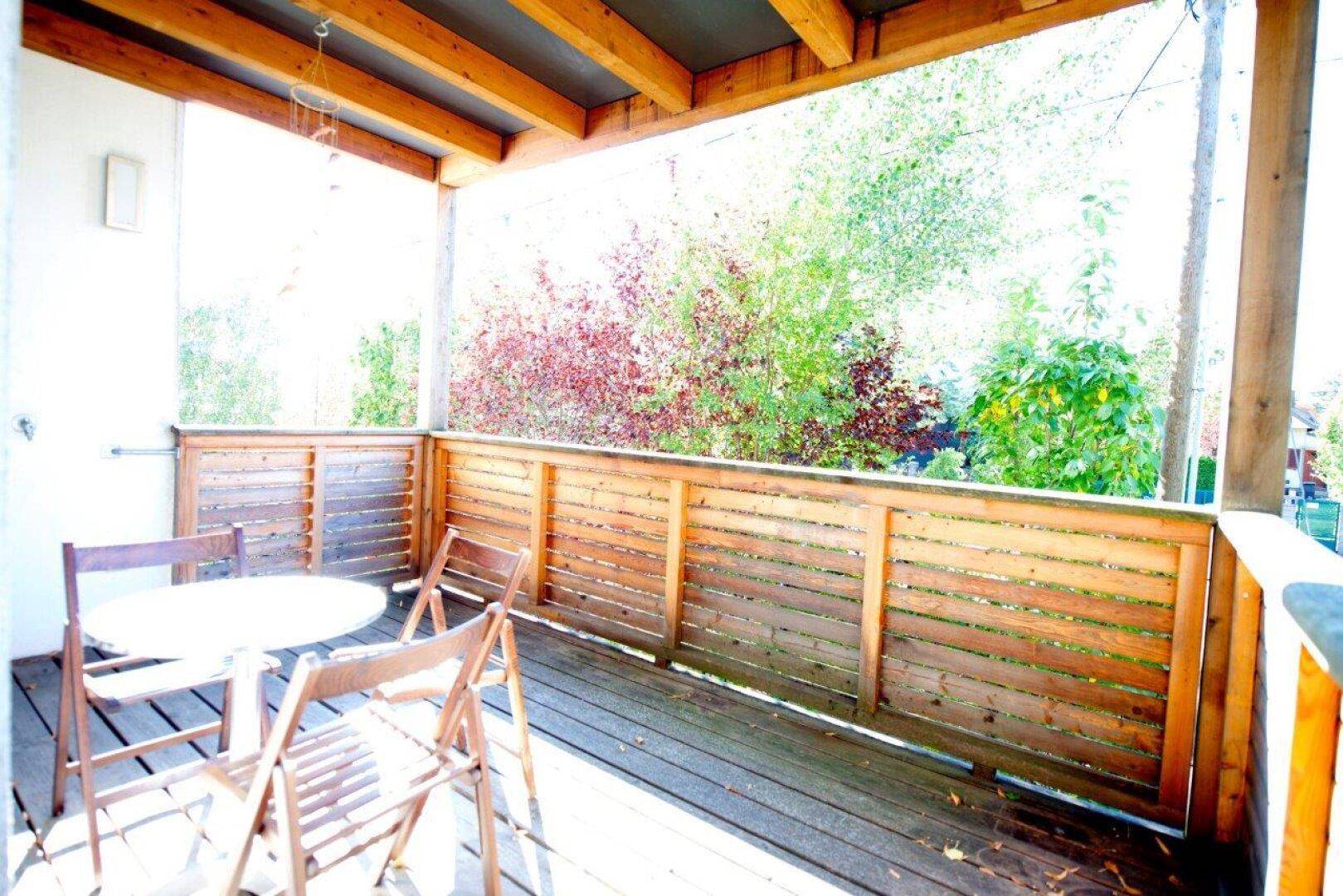 Überdachter Balkon mit Holzboden und Sitzgelegenheiten, umgeben von Bäumen.