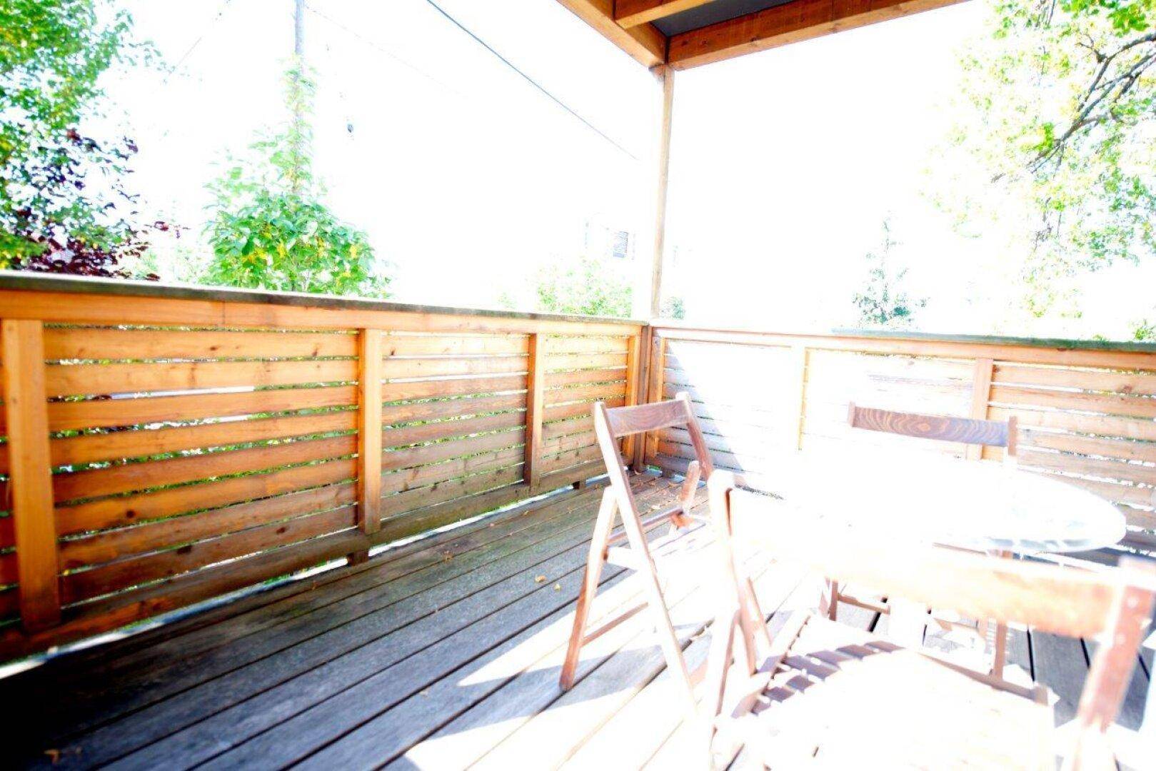 Sonniger Balkon mit Holzboden und Sitzgelegenheiten, umgeben von Bäumen.