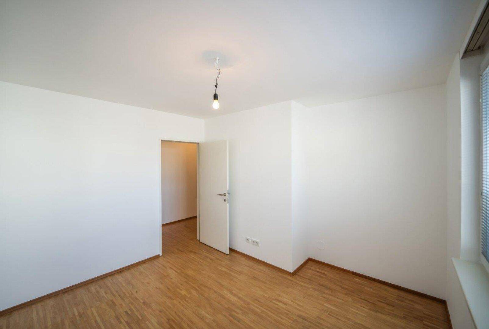 Leeres Zimmer mit Parkettboden, weißen Wänden und natürlichem Lichteinfall durch das Fenster.
