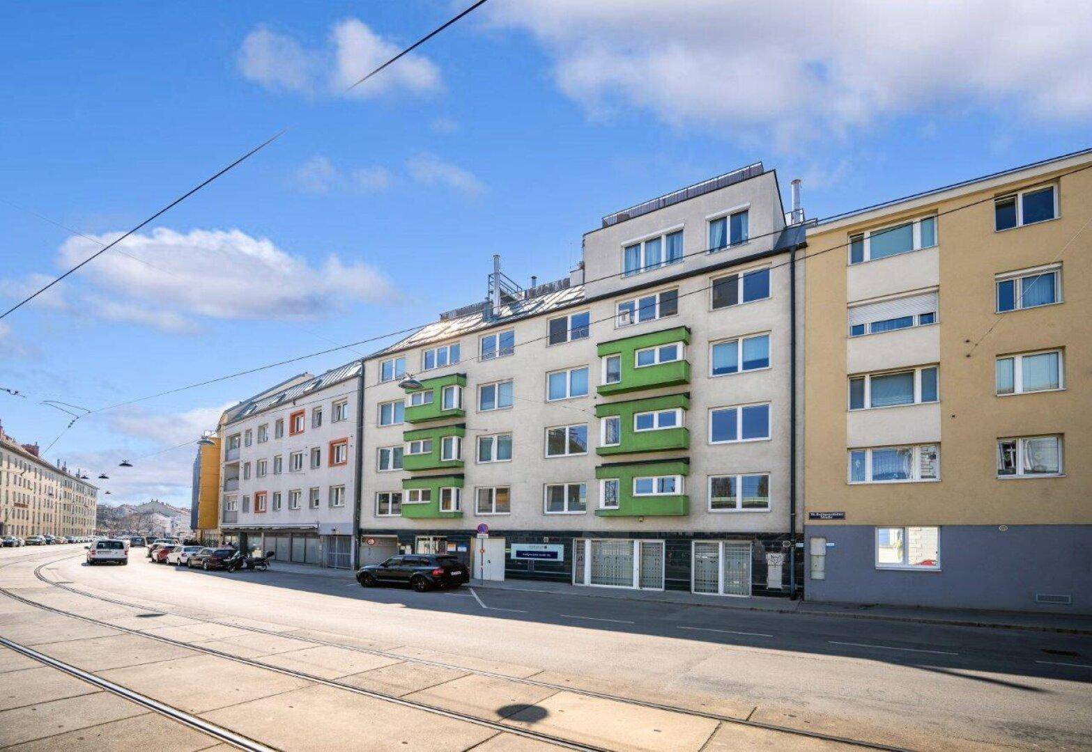 Mehrfamilienhaus mit grünen Balkonen an einer belebten Straße unter blauem Himmel.