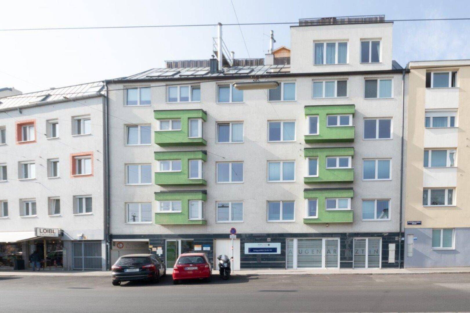 Mehrfamilienhaus mit moderner Fassade, grünen Balkonen und Geschäften im Erdgeschoss.