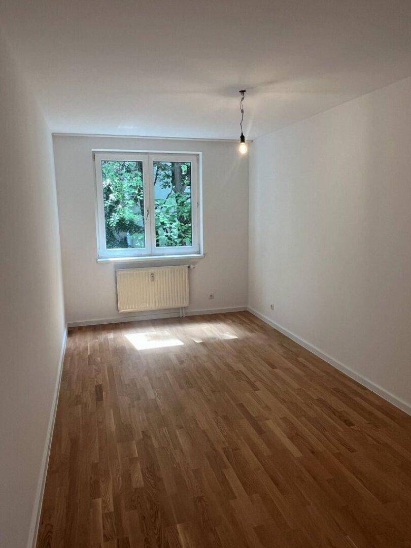 Helles Schlafzimmer mit Parkettboden und Fenster, das viel Tageslicht hereinlässt und einen grünen Blick bietet.