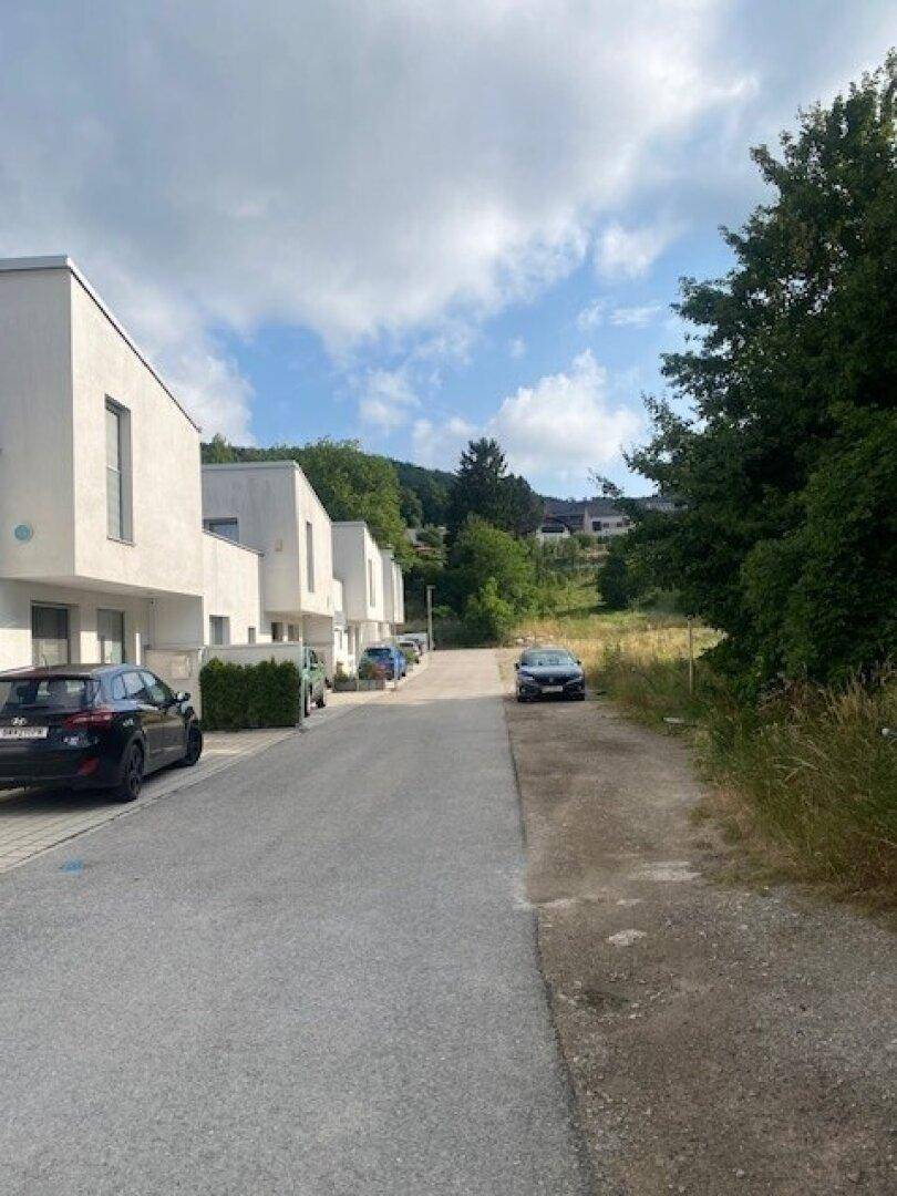 Reihen moderner Häuser entlang einer Straße mit parkenden Autos und grüner Umgebung.