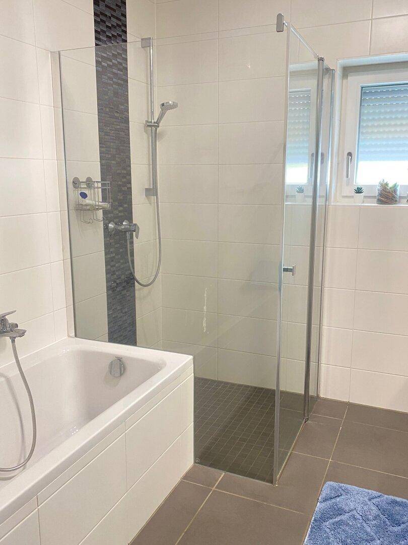 Modernes Badezimmer mit Badewanne, Duschkabine und Fenster, gefliest in Weiß und Grau.