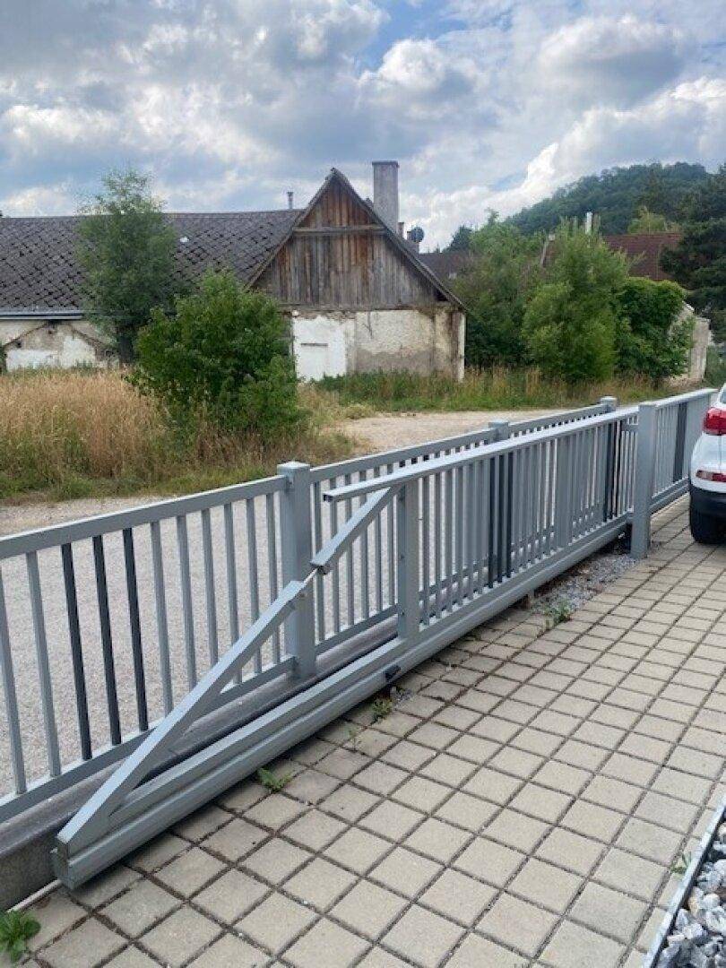 Grauer Schiebetorzaun an einer gepflasterten Einfahrt mit Blick auf ein altes Gebäude.
