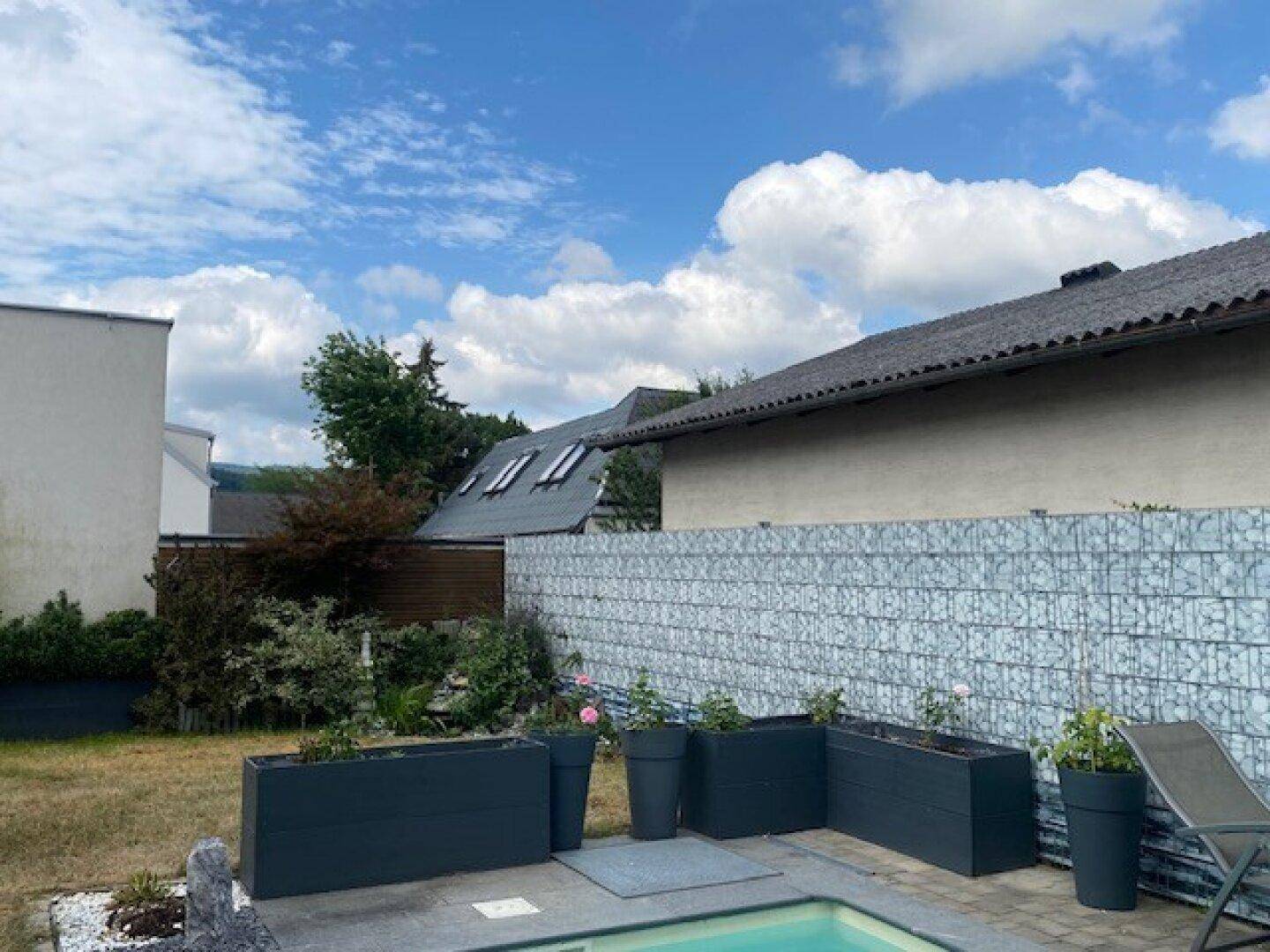 Moderner Außenpool mit Gabionenwand, umgeben von Pflanzen und einer Liegefläche.