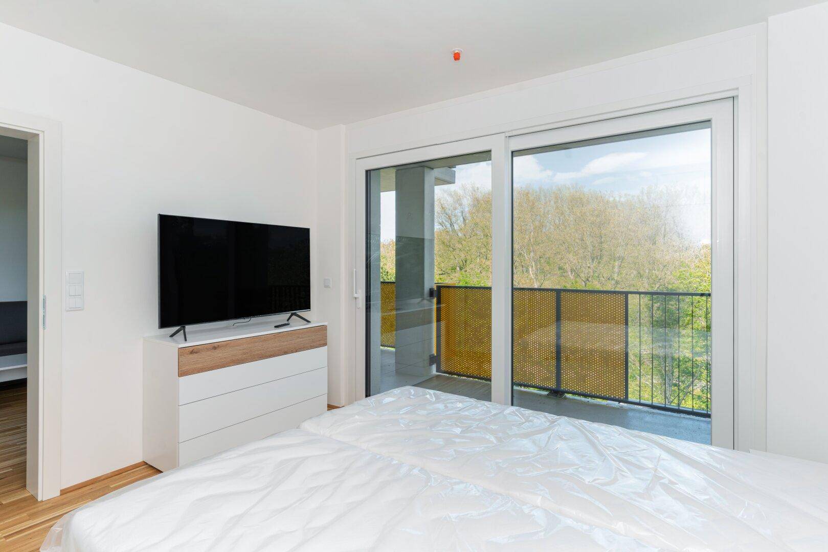 Schlafzimmer mit Fernseher, Kommode und großem Fenster mit Blick ins Grüne.