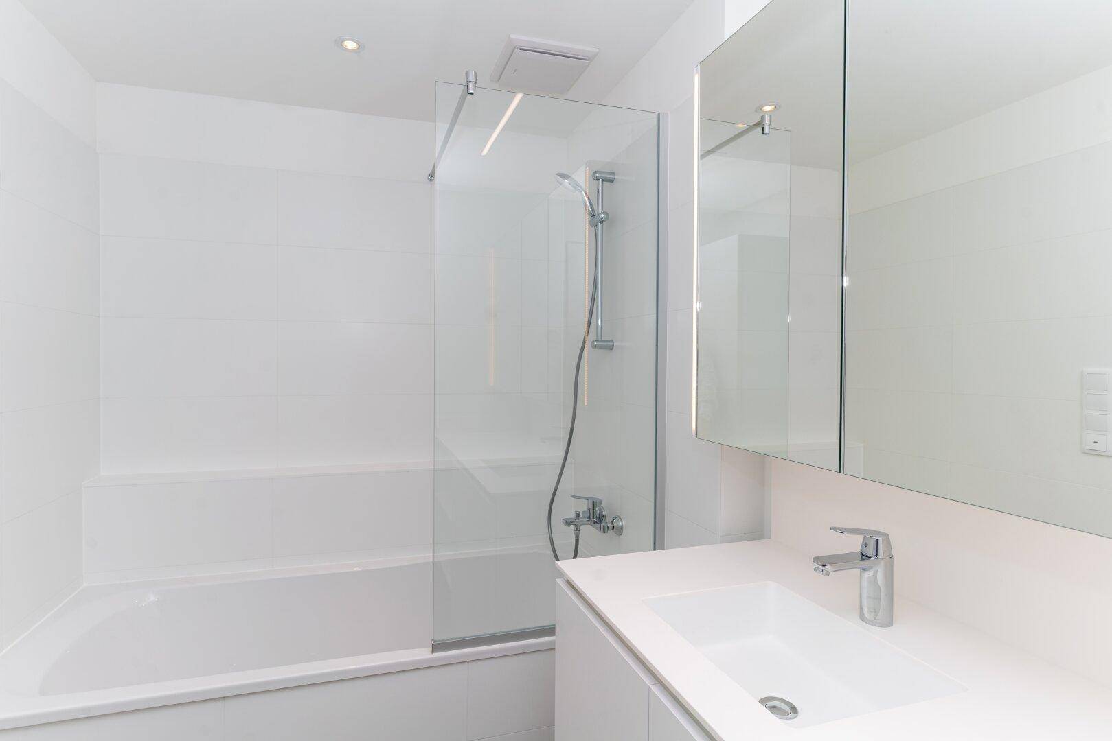 Helles Badezimmer mit Badewanne, Glasduschwand und integriertem Waschbecken in minimalistischem Stil.