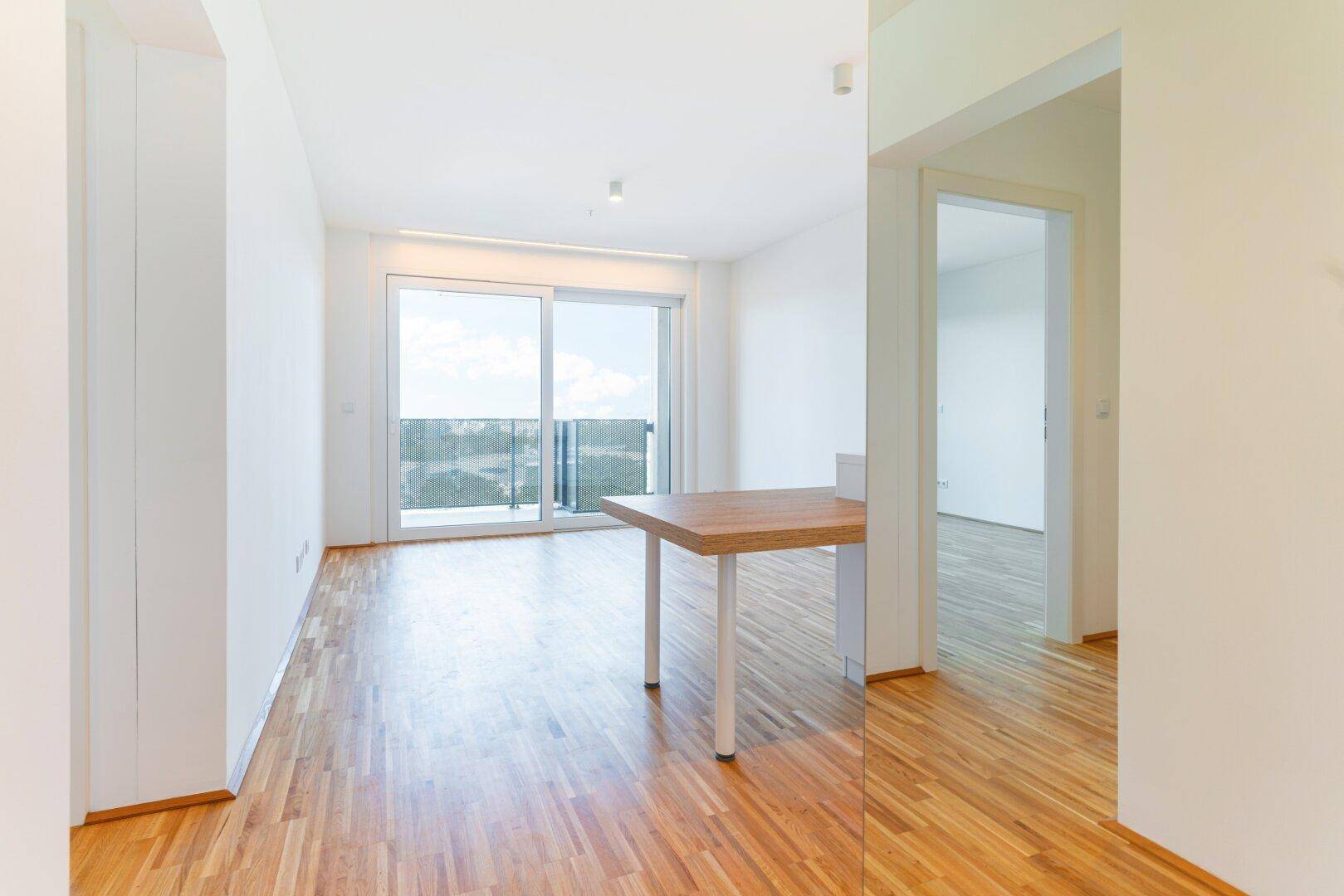 Heller offener Wohnbereich mit Holzboden, großen Fenstern und Zugang zu einem Balkon.