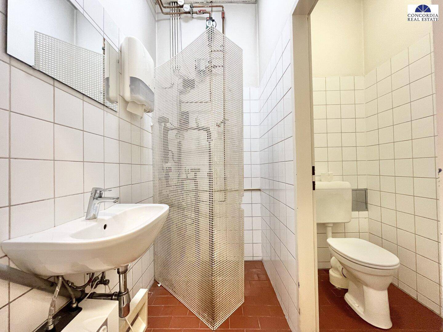 Sanitärbereich mit Waschbecken, Toilette und weißen Fliesen, abgetrennt durch eine perforierte Metallwand.