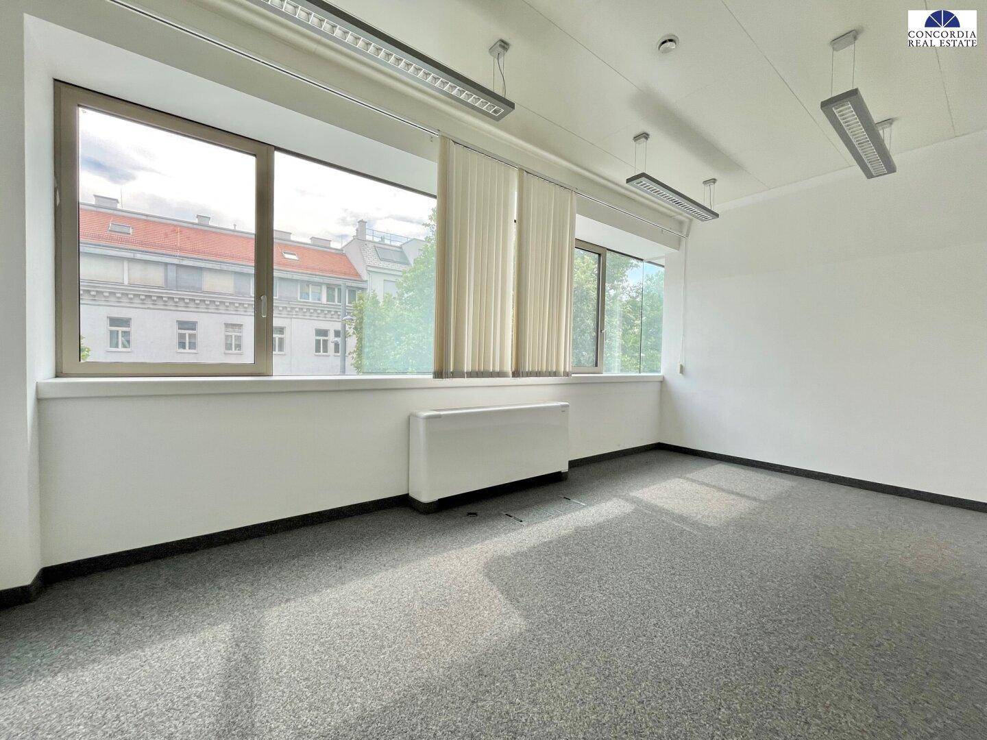Heller Büroraum mit Teppichboden, breiten Fenstern und Blick auf die umliegenden Gebäude.