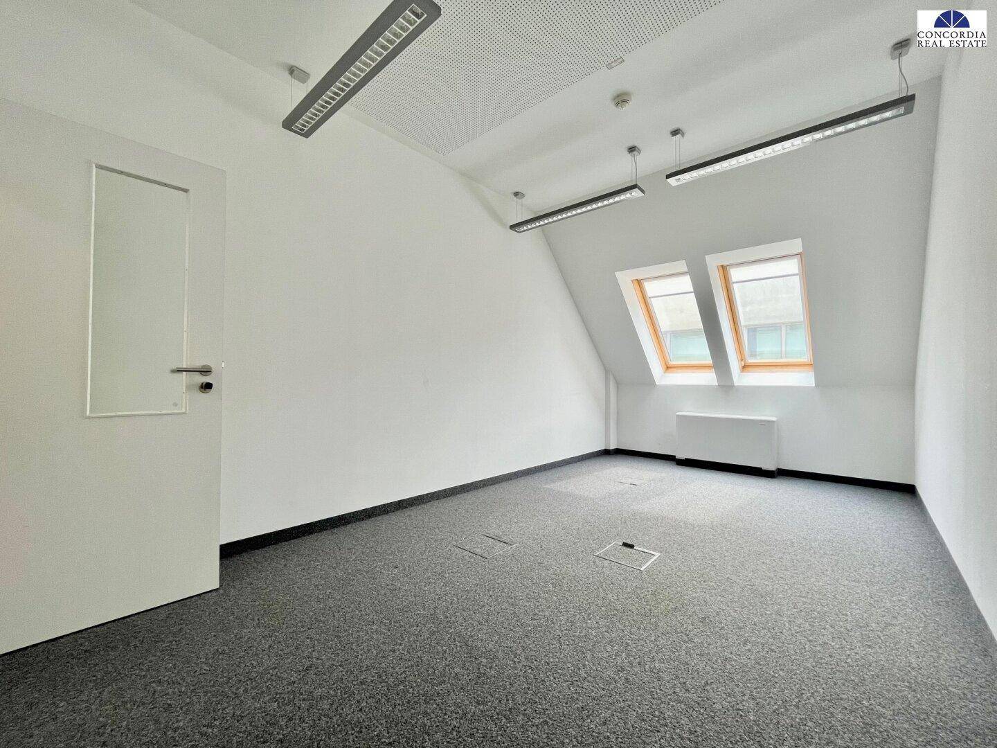 Leerer Büroraum im Dachgeschoss mit Teppichboden, Dachschräge und zwei Dachfenstern.