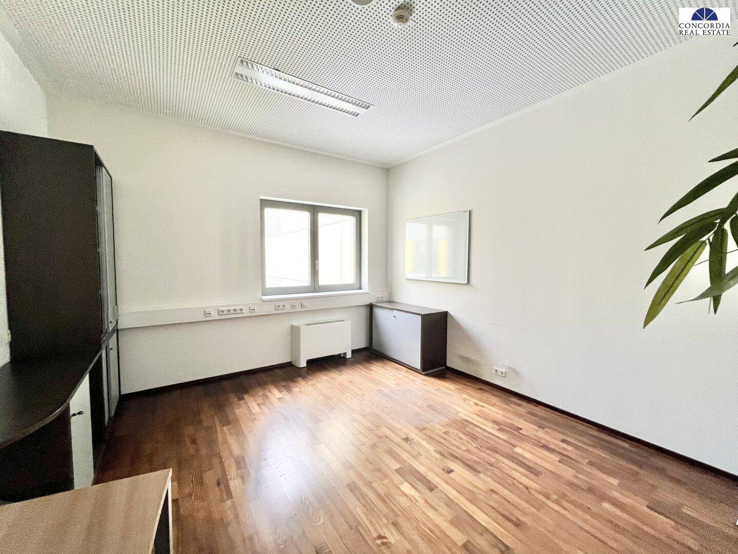 Leeres Büro mit Holzboden, weißen Wänden und einem großen Fenster.