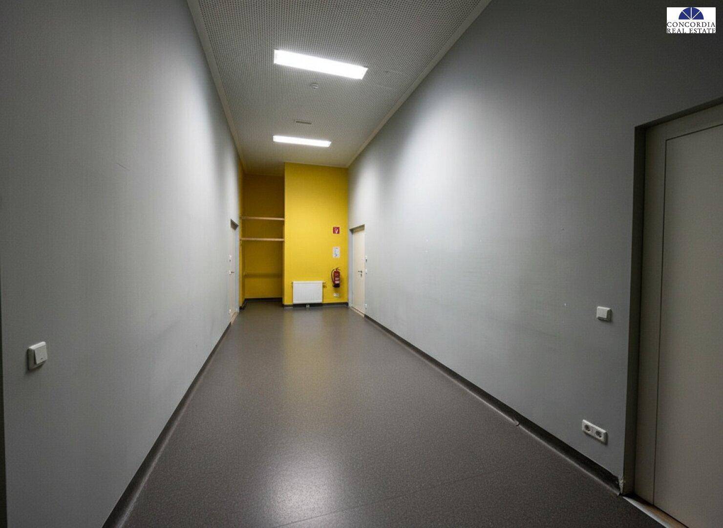 Langer, heller Flur mit grauen Wänden und einem gelben Akzentbereich am Ende.
