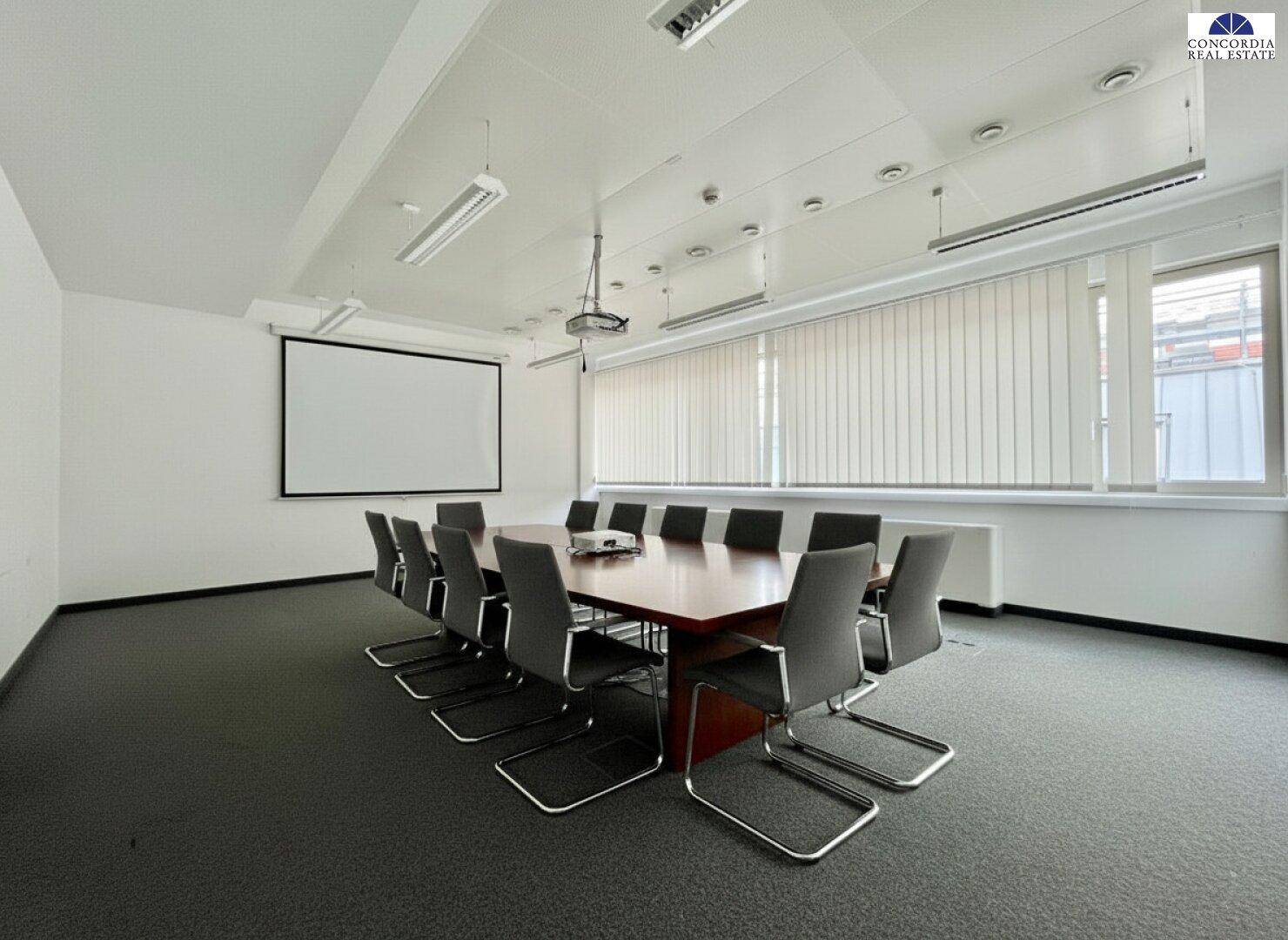 Geräumiger Konferenzraum mit großem Tisch, Stühlen, Beamer und Leinwand für Präsentationen.