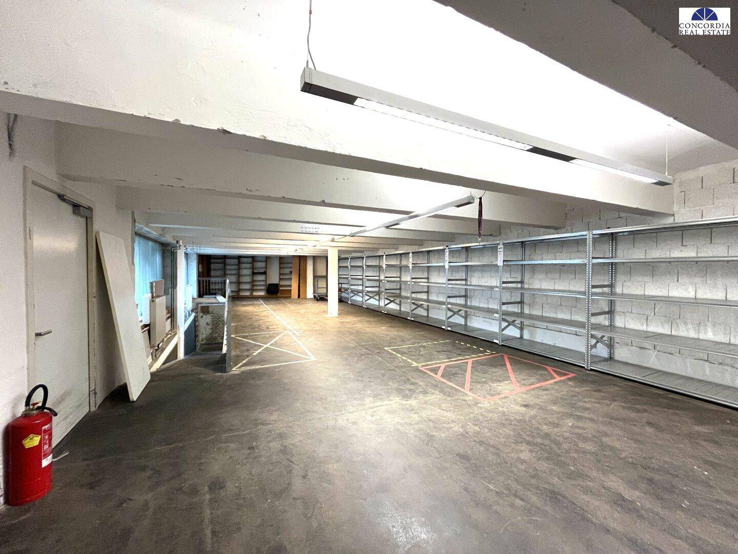 Großer leerer Lagerraum mit Betonboden, weißen Wänden und hohen Metallregalen an der Seite.