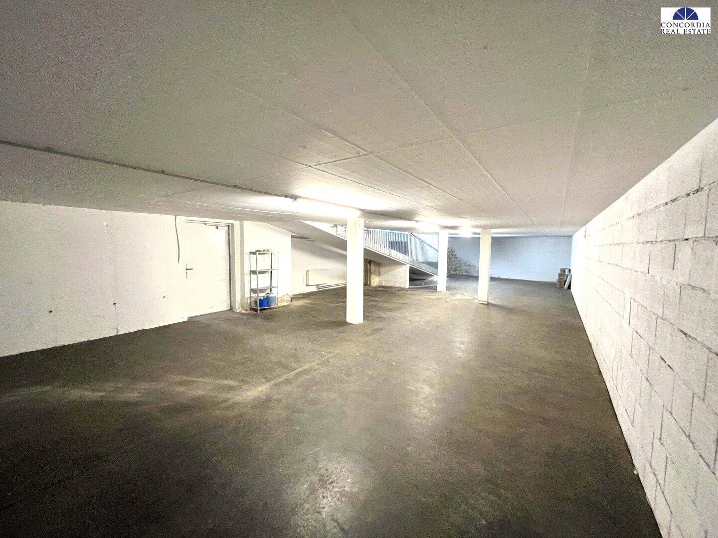 Geräumiger Lagerraum mit Betonboden, weißen Säulen und einer Treppe im hinteren Bereich.