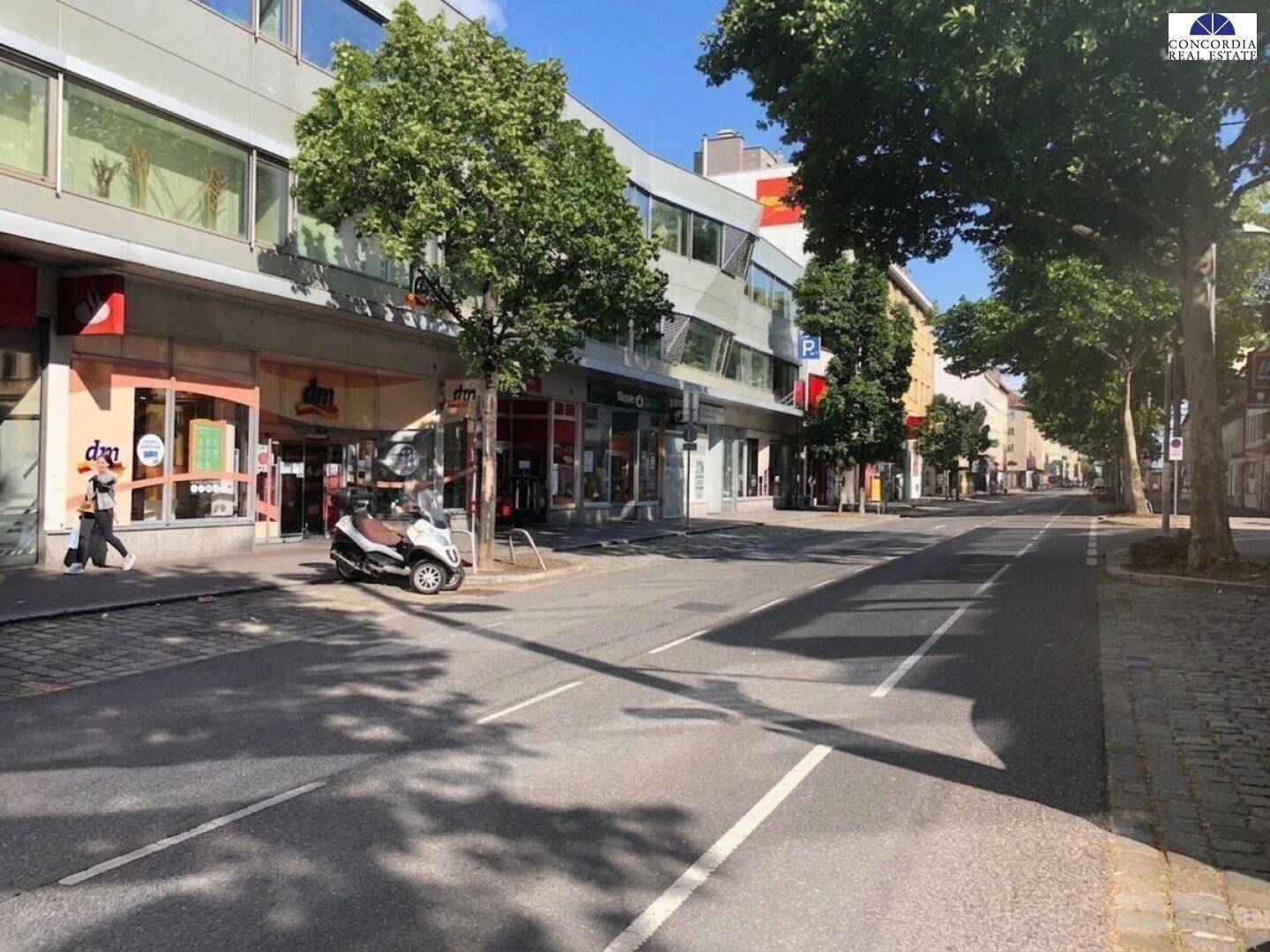 Belebtes Stadtbild mit Geschäften, Bäumen und einer Straße unter blauem Himmel.