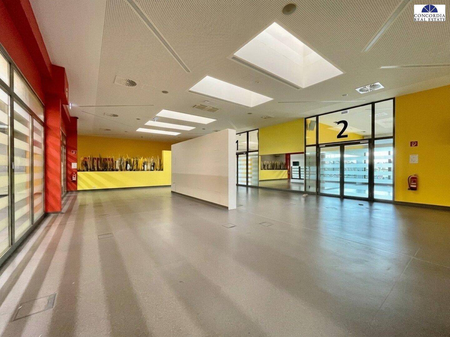 Geräumiger Saal mit großen Glasfronten und gelben Wänden, ideal für Veranstaltungen.
