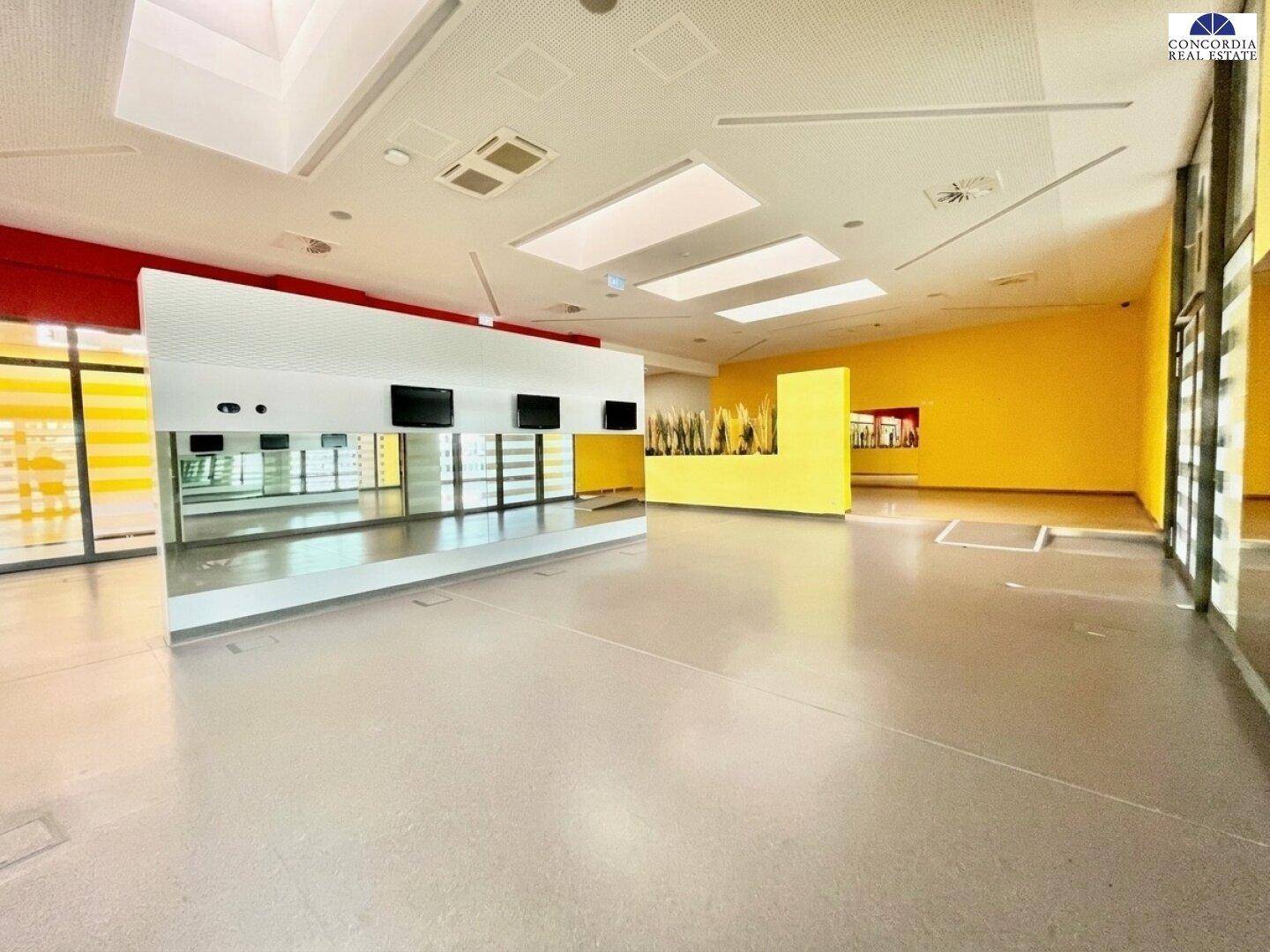 Großer, leerer Saal mit Spiegelfront, gelben Wänden und modernen Deckenleuchten.
