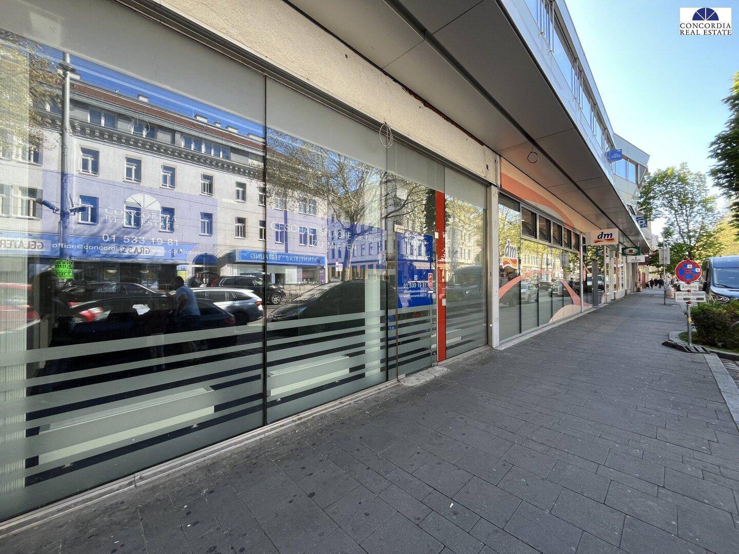 Lange Geschäftsfassade entlang eines belebten Bürgersteigs mit reflektierenden Schaufenstern und angrenzenden Geschäften.