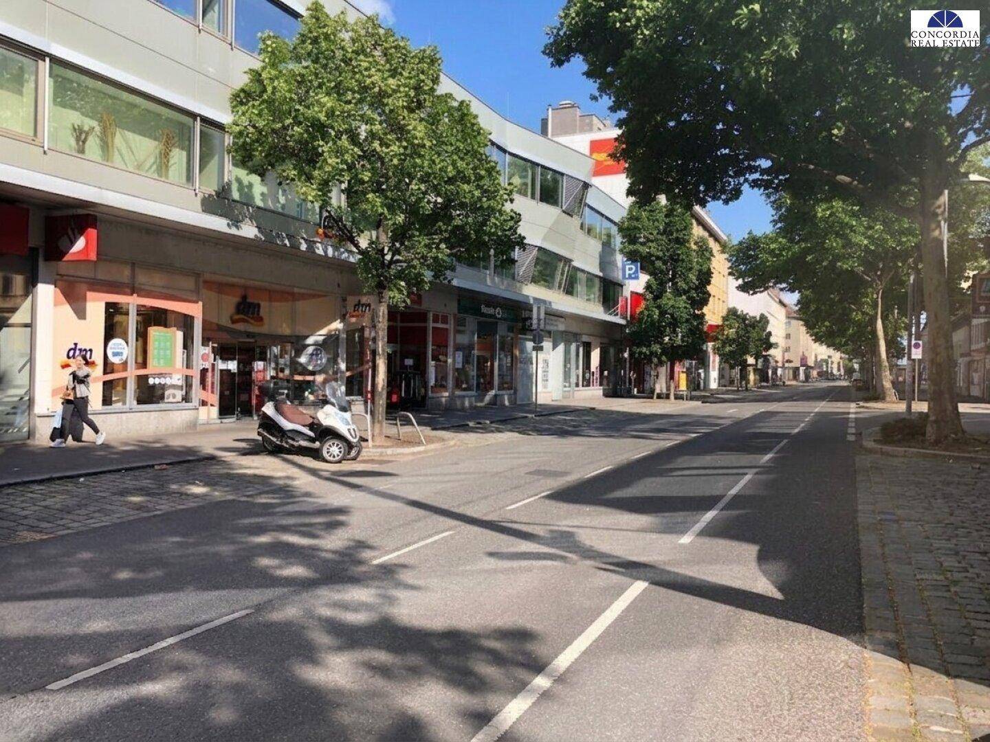 Belebtes Stadtbild mit Geschäftsgebäuden, Bäumen und einer Straße mit parkenden Fahrzeugen an einem sonnigen Tag.