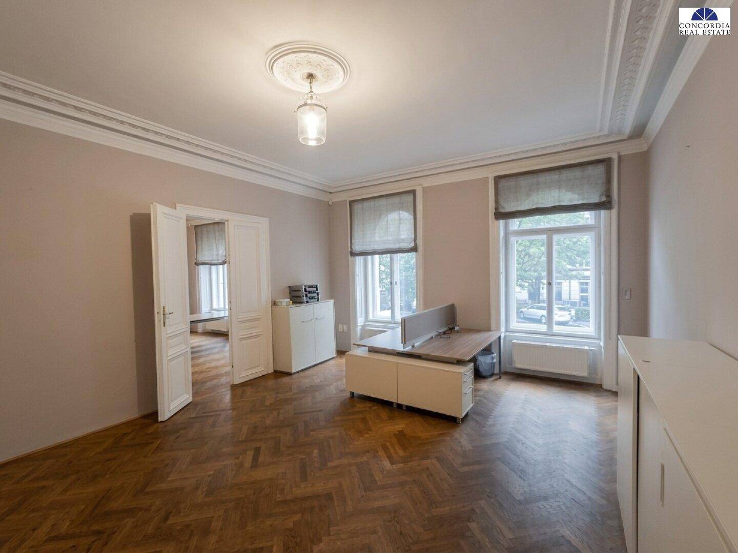 Großes Büro mit Fischgrätparkett, hohen Decken und Blick auf die Straße durch die Fenster.