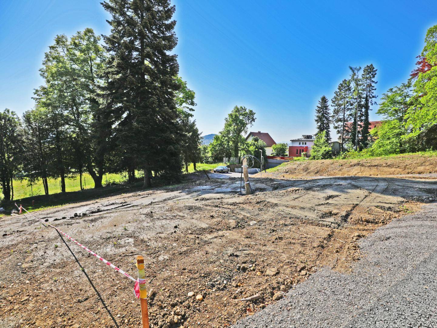 Offenes Baugrundstück mit Schotter und Erde, umgeben von Bäumen und mit einem Fahrzeug in der Ferne.