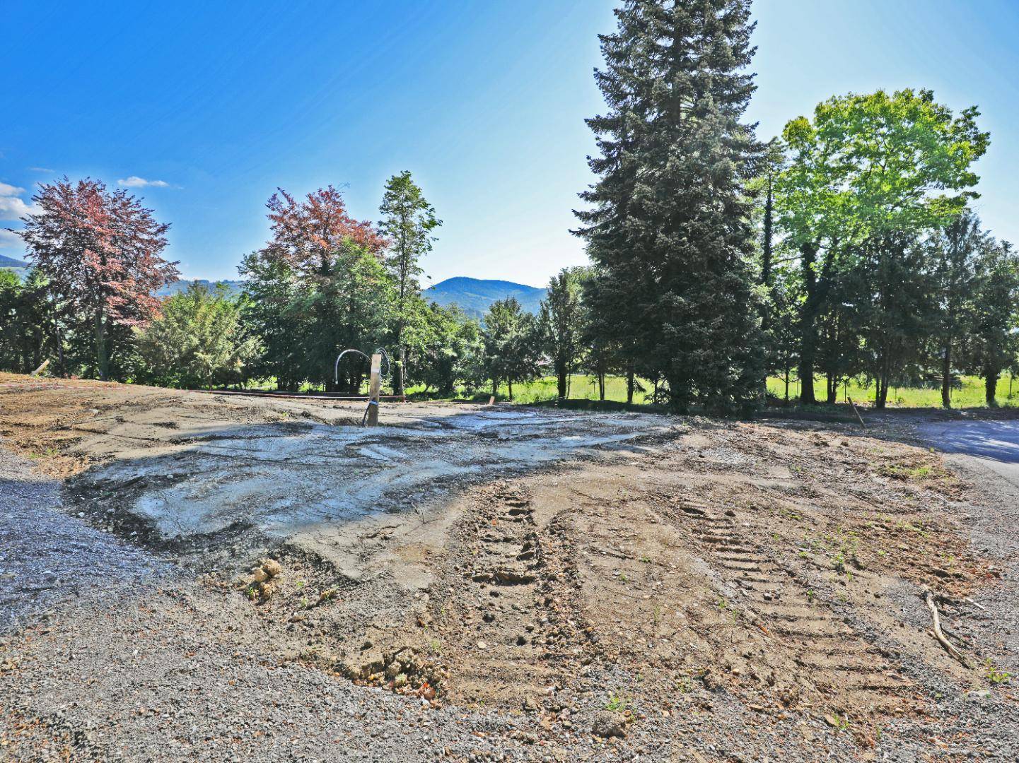 Freie Baufläche mit Schotter und Erde, umgeben von Bäumen und mit Blick auf ferne Berge.