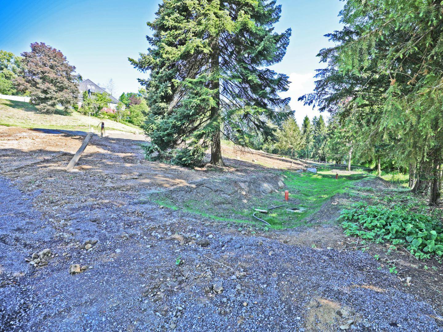 Baugrundstück mit sichtbaren Versorgungsleitungen in einem Graben, umgeben von Bäumen und Grünflächen.