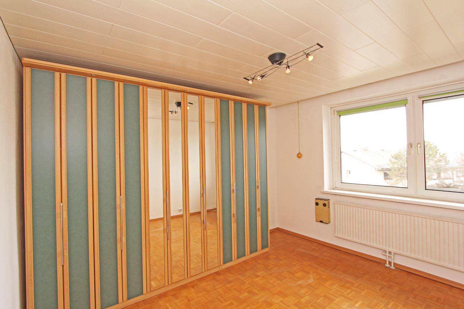 Schlafzimmer mit großem Einbauschrank, Parkettboden und Fenster mit Blick nach draußen.