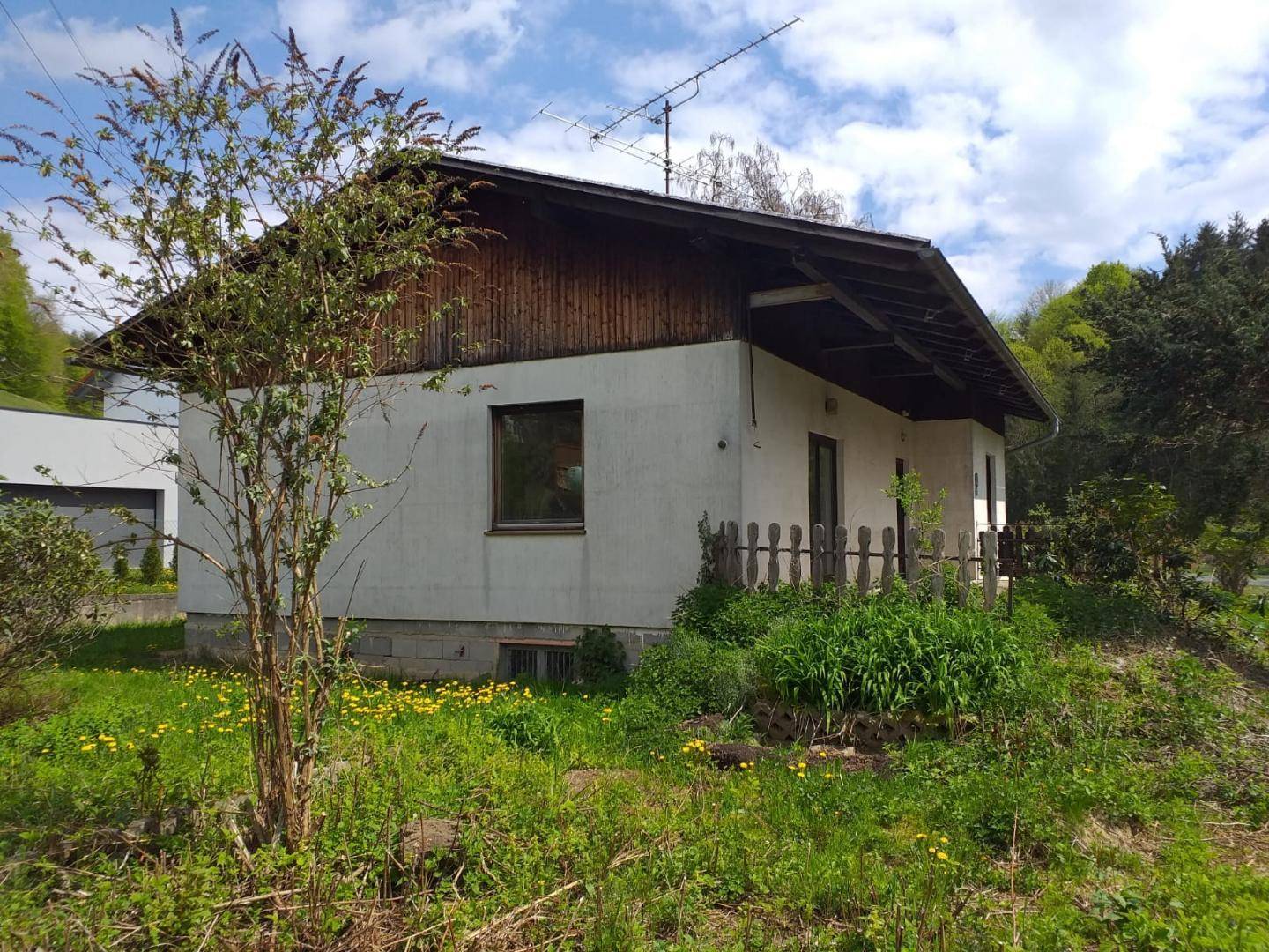 Seitenansicht des Hauses mit Holzelementen unter dem Dach und einfacher Umzäunung im Grünen.