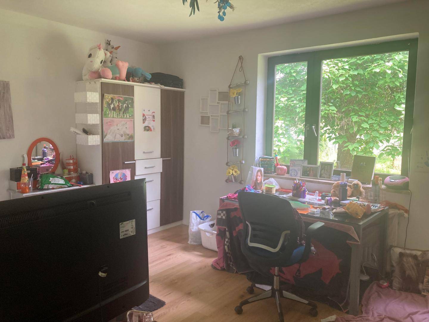 Helles Schlafzimmer mit Schreibtisch, Kleiderschrank und großem Fenster mit Grünblick.