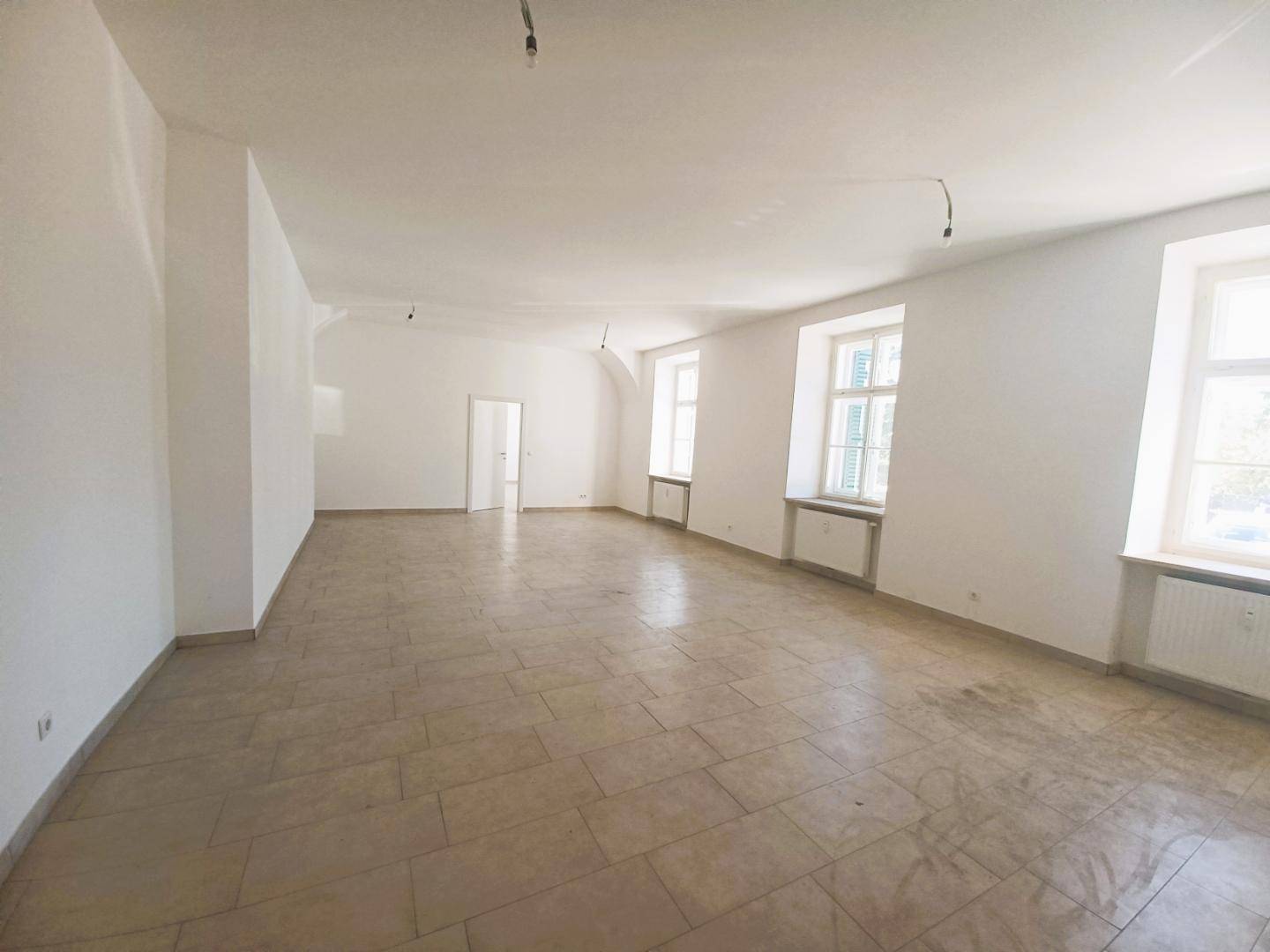 Heller, leerer Raum mit Fliesenboden, hohen Fenstern und weiß gestrichenen Wänden.