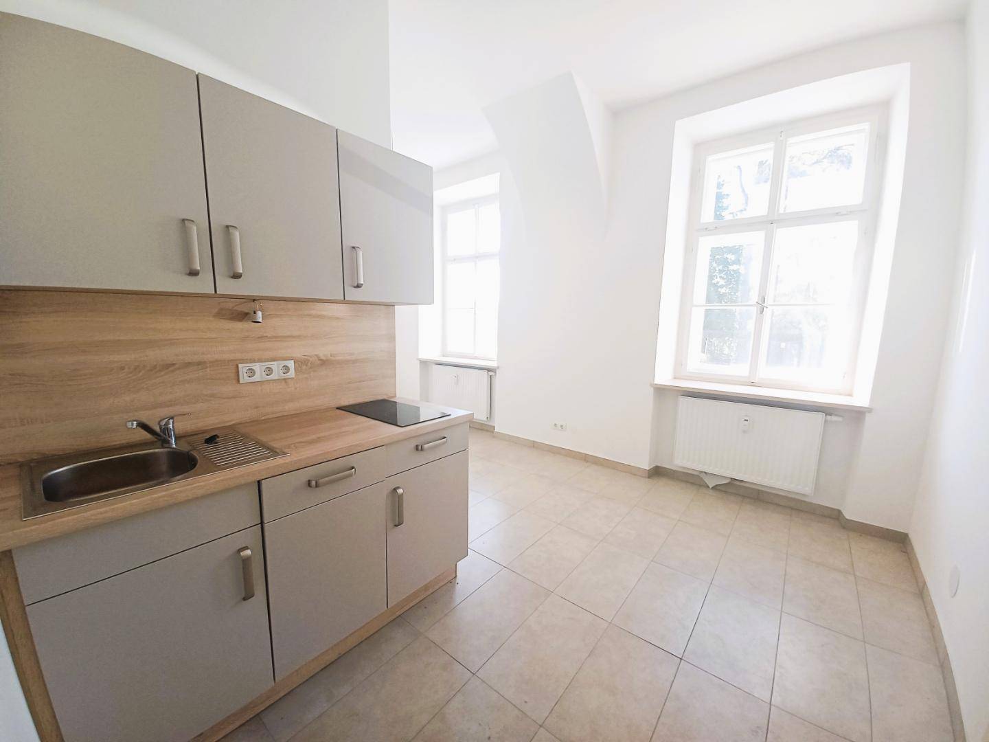 Kompakte Küchenzeile mit hellgrauen Schränken, Holzarbeitsplatte und integriertem Kochfeld in einem hellen Raum.