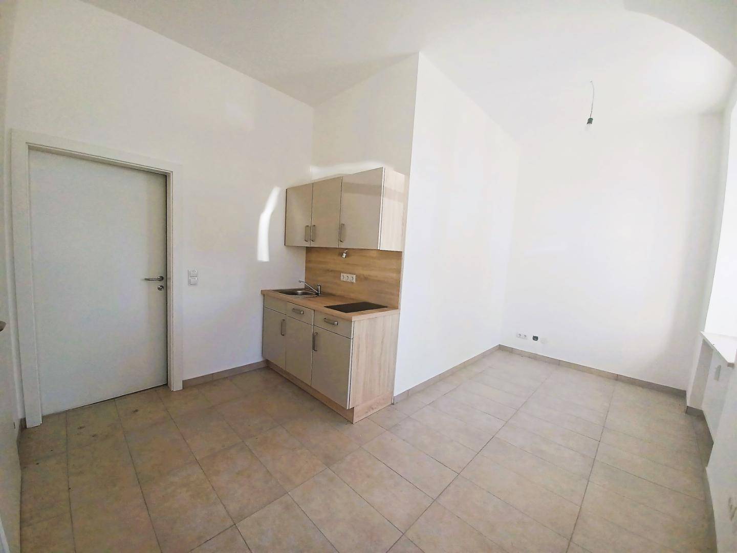 Kleine Küchenzeile mit Oberschränken und Arbeitsplatte, daneben eine weiße Tür in einem gefliesten Raum.